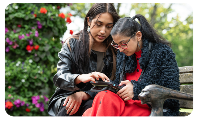 Sufina uses a Talk Pad device from Smartbox to communicate. She is sitting outside on a park bench with her sister.